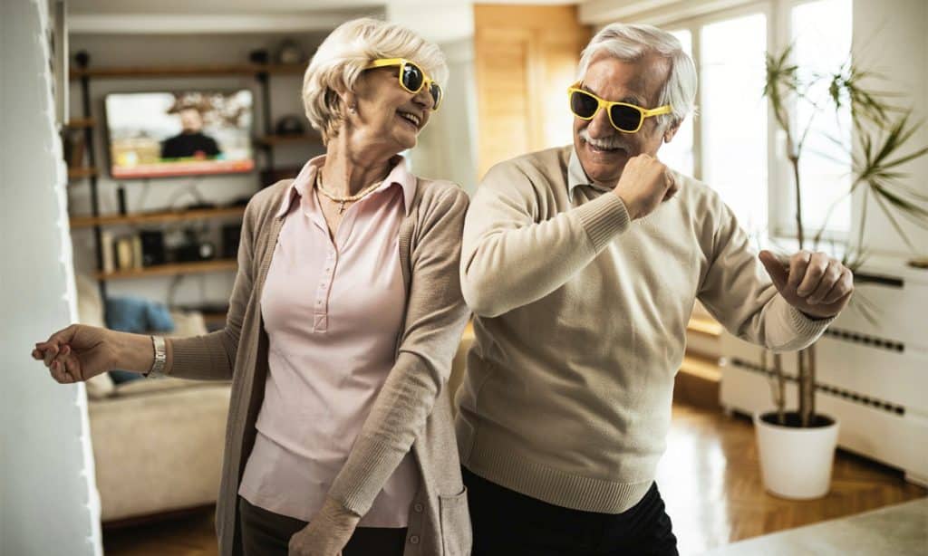 Un couple de senior heureux qui danse dans le salon de leur logement avec des lunettes jaunes