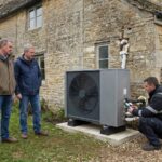 Deux proprietaires et un technicien devant une pompe a chaleur air-eau installee contre une maison ancienne en renovation d'ampleur