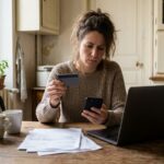 Une femme a la table de sa cuisine tient une carte bancaire et son telephone devant des factures papier et un ordinateur portable