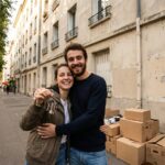 Jeune couple souriant tenant les cles de leur premier logement devant un immeuble avec des cartons de demenagement