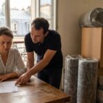 Un couple remplit un dossier d'aide a la renovation energetique dans son appartement, rouleaux d'isolation et systeme de ventilation poses au sol