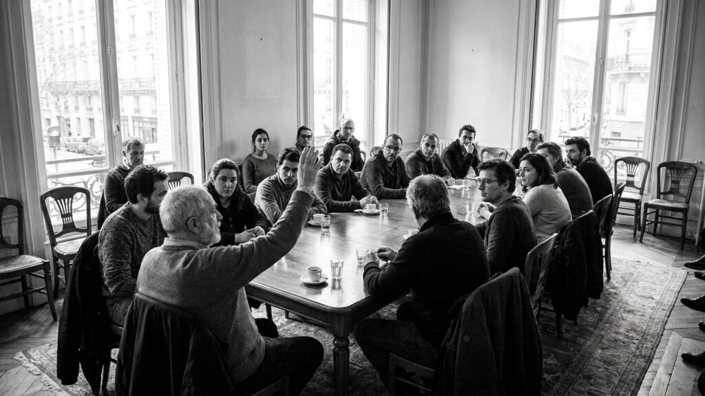 Reunion d'assemblee generale de copropriete autour d'une table, un participant leve la main pour voter