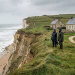Deux promeneurs en cires observent la mer depuis une falaise erodee du littoral francais, avec des maisons de bord de mer en arriere-plan