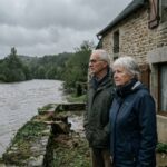 Un couple de proprietaires observe une riviere en crue devant leur residence secondaire en pierre
